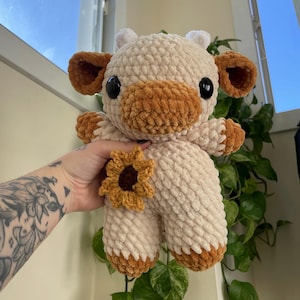 Puede incluir: Un juguete de peluche de vaca tejido a crochet con una nariz marr&oacute;n y un detalle de girasol. La vaca es blanca con detalles marrones y tiene grandes ojos negros.