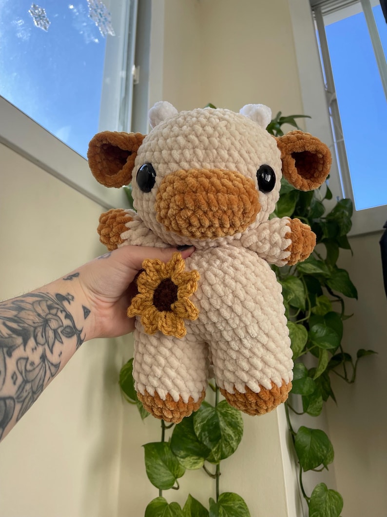 Puede incluir: Un juguete de peluche de vaca tejido a crochet con una nariz marr&oacute;n y un detalle de girasol. La vaca es blanca con detalles marrones y tiene grandes ojos negros.