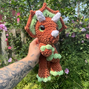 May include: A brown crocheted triceratops plush toy with green and white accents. The toy has large, black eyes and a white horn. The toy is being held in a hand.