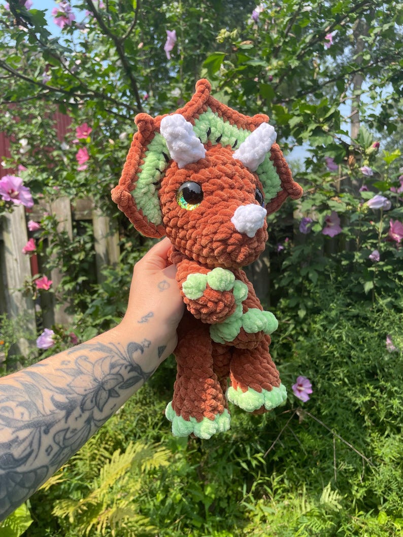 May include: A brown crocheted triceratops plush toy with green and white accents. The toy has large, black eyes and a white horn. The toy is being held in a hand.