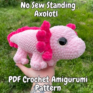 May include: A pink and fuchsia crochet axolotl amigurumi. The plush toy has large, sparkly black eyes and a frilly, textured exterior. The text "No Sew Standing Axolotl" and "PDF Crochet Amigurumi Pattern" are displayed.