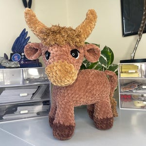 以下が含まれることがあります： 茶色とベージュの毛糸で作られたハイランドカウのぬいぐるみ。質感のあるふわふわした体、大きな暗い色の目、2本の湾曲した角、頭の上のボサボサした毛の房が特徴です。ひづめは濃い茶色をしています。