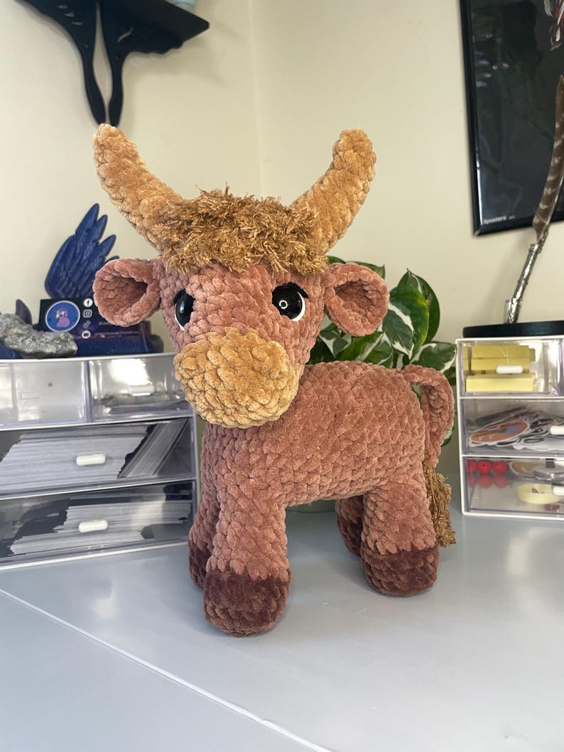 以下が含まれることがあります： 茶色とベージュの毛糸で作られたハイランドカウのぬいぐるみ。質感のあるふわふわした体、大きな暗い色の目、2本の湾曲した角、頭の上のボサボサした毛の房が特徴です。ひづめは濃い茶色をしています。