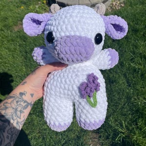 Puede incluir: Una vaca de peluche blanca tejida a crochet con orejas, pezu&ntilde;as y nariz moradas. La vaca sostiene una flor morada con hojas verdes.