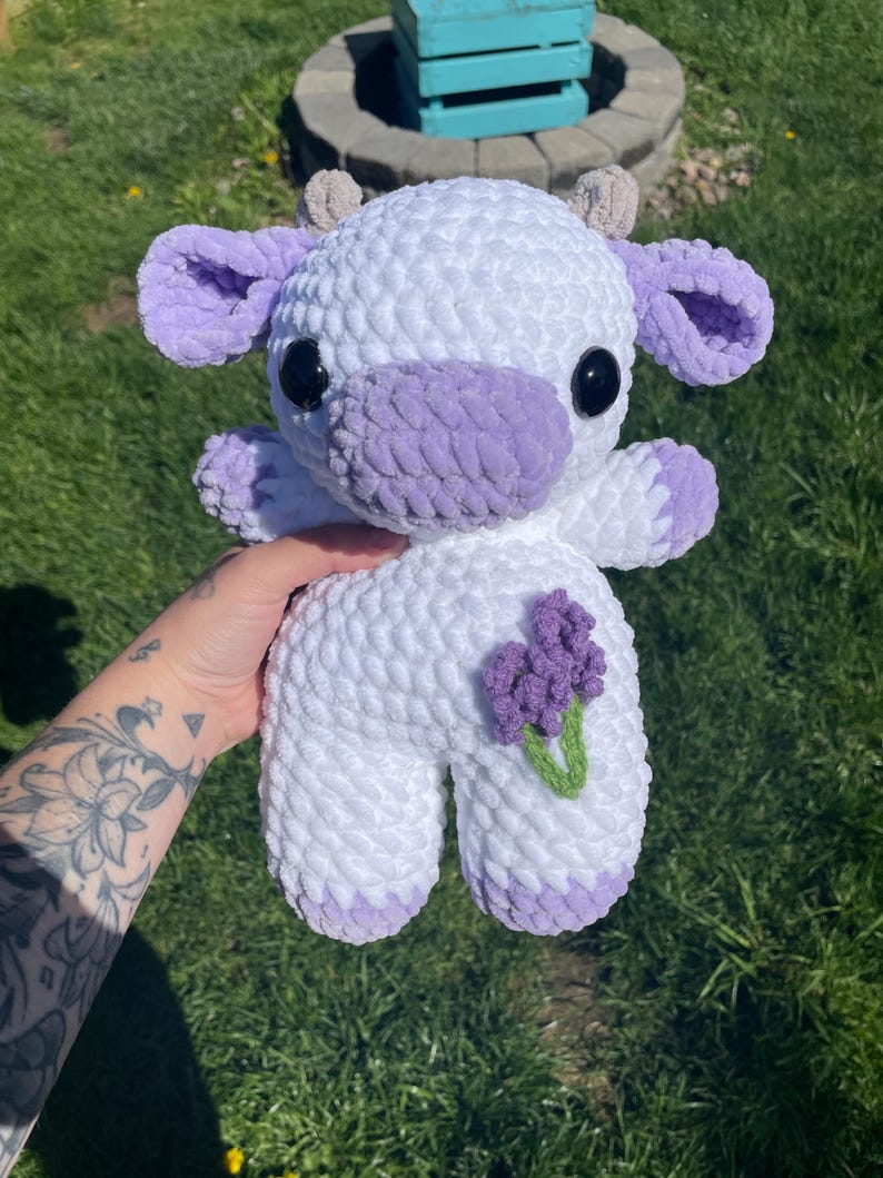 Puede incluir: Una vaca de peluche blanca tejida a crochet con orejas, pezu&ntilde;as y nariz moradas. La vaca sostiene una flor morada con hojas verdes.