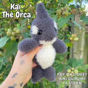 May include: A handmade gray and white amigurumi orca plush toy. The plush has black eyes and is labeled "Kai The Orca." The image also includes text that reads "PDF Crochet Amigurumi Pattern."