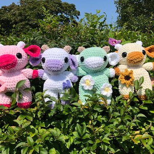 Puede incluir: Cuatro peluches de vaca de crochet con diferentes colores de pelo y detalles de flores. Las vacas son rosa, blanca con detalles morados, verde y blanca con detalles marrones. Las flores son rosa, blanca y amarilla.