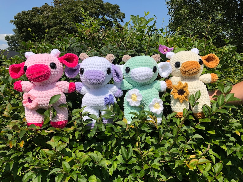 Puede incluir: Cuatro peluches de vaca de crochet con diferentes colores de pelo y detalles de flores. Las vacas son rosa, blanca con detalles morados, verde y blanca con detalles marrones. Las flores son rosa, blanca y amarilla.