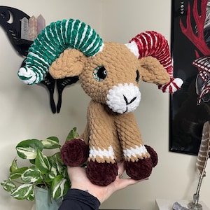 May include: A handmade crochet ram plushie with brown, white, green, and red accents. The ram has large, colorful horns, a white face, and brown legs with white and burgundy details. The plushie is held in a person's hand.