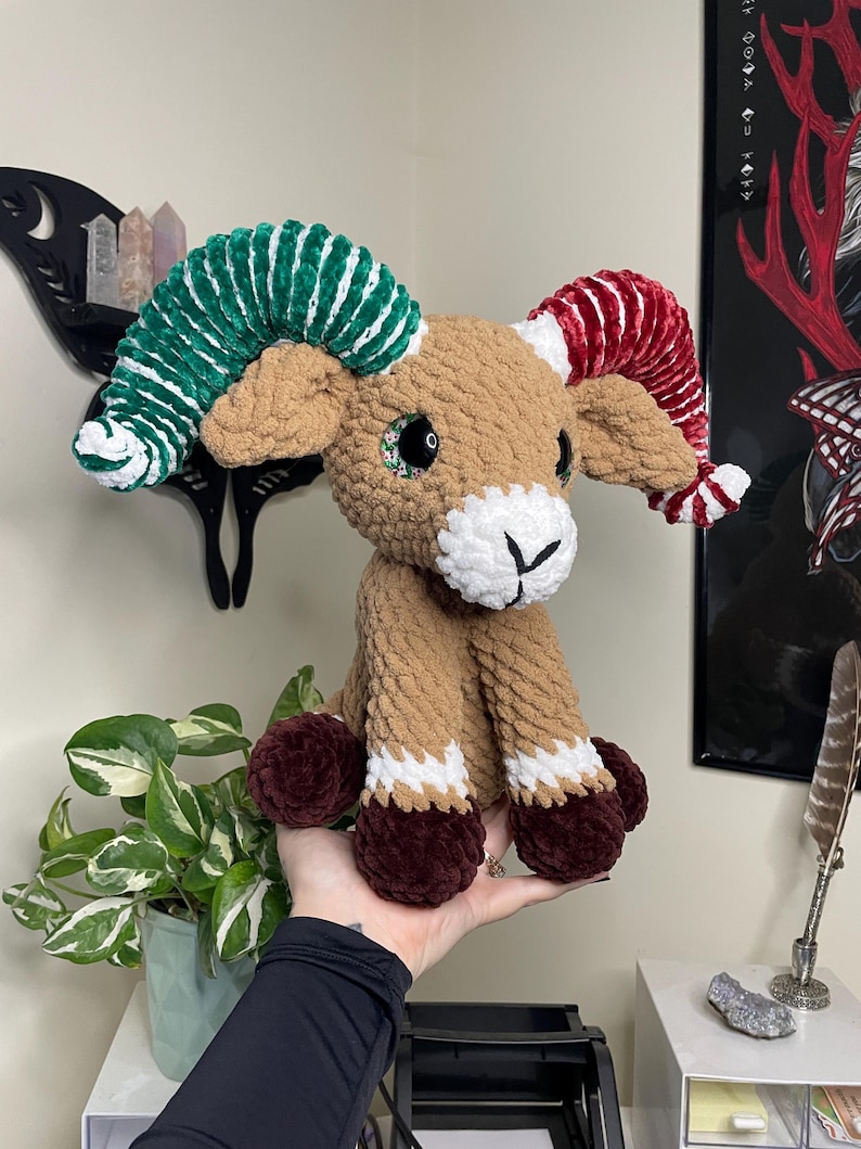 May include: A handmade crochet ram plushie with brown, white, green, and red accents. The ram has large, colorful horns, a white face, and brown legs with white and burgundy details. The plushie is held in a person's hand.