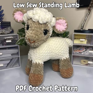 May include: A handmade crochet lamb with a fluffy white body, tan face and legs, and pink-lined ears. The text "Low Sew Standing Lamb" and "PDF Crochet Pattern" are displayed above and below the lamb.