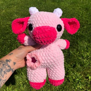 Puede incluir: Un juguete de peluche de vaca de crochet rosa con cuernos blancos y detalles rosas. El juguete tiene ojos negros y una nariz rosa.