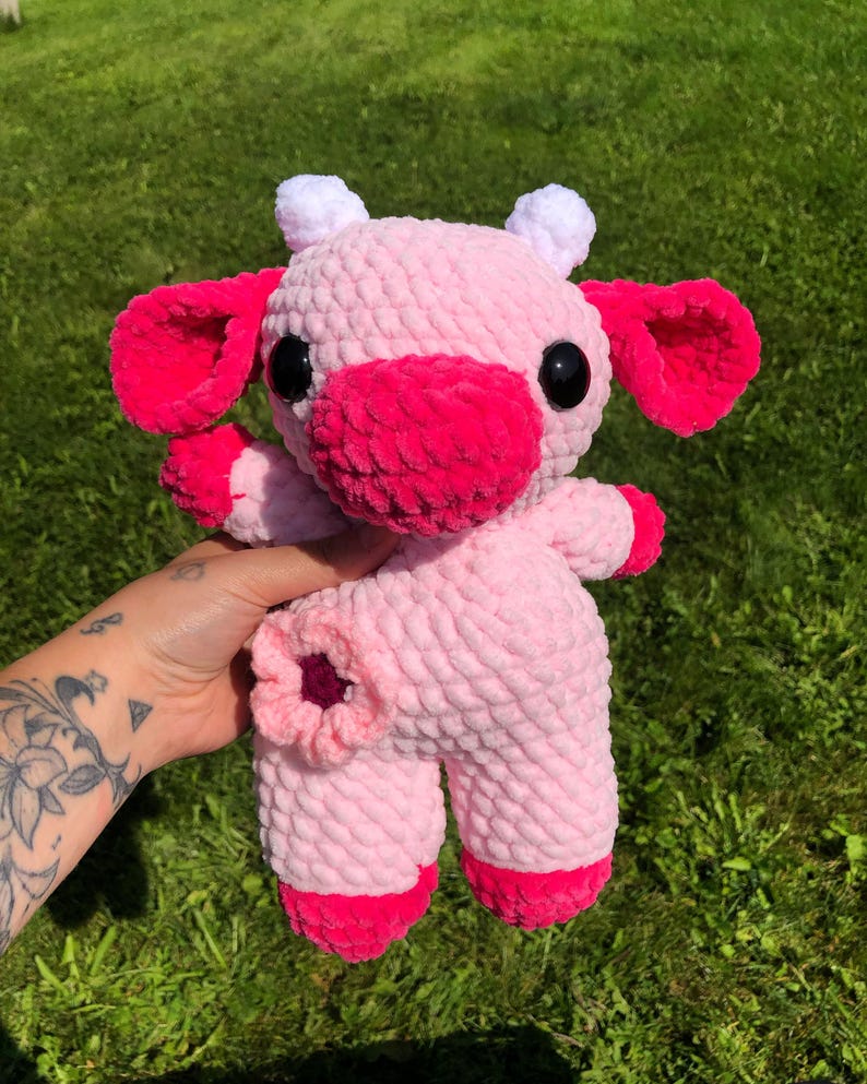 Puede incluir: Un juguete de peluche de vaca de crochet rosa con cuernos blancos y detalles rosas. El juguete tiene ojos negros y una nariz rosa.