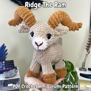 Może przedstawiać: Szydełkowy amigurumi baran z brązowymi i beżowymi detalami. Baran ma duże, zakręcone rogi, czarne oczy i biały pysk. Tekst "Ridge The Ram" znajduje się nad baranem, a "PDF Crochet Amigurumi Pattern" pod nim.