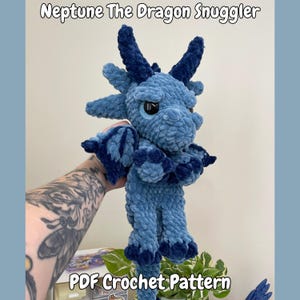 Könnte beinhalten: Ein handgefertigtes, hellblaues Häkeldrachen-Spielzeug mit dunkelblauen Akzenten an Flügeln, Hörnern und Füßen. Der Drache hat große, schwarze Augen und ist mit "Neptune The Dragon Snuggler" und "PDF Crochet Pattern" beschriftet.