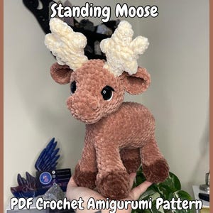 Könnte beinhalten: Ein stehendes Elch-Amigurumi aus Plüsch in Braun- und Cremetönen. Der Elch hat große, schwarze Augen und cremefarbene Geweihe. Der Text "Standing Moose" und "PDF Crochet Amigurumi Pattern" sind oberhalb und unterhalb des Elchs zu sehen.