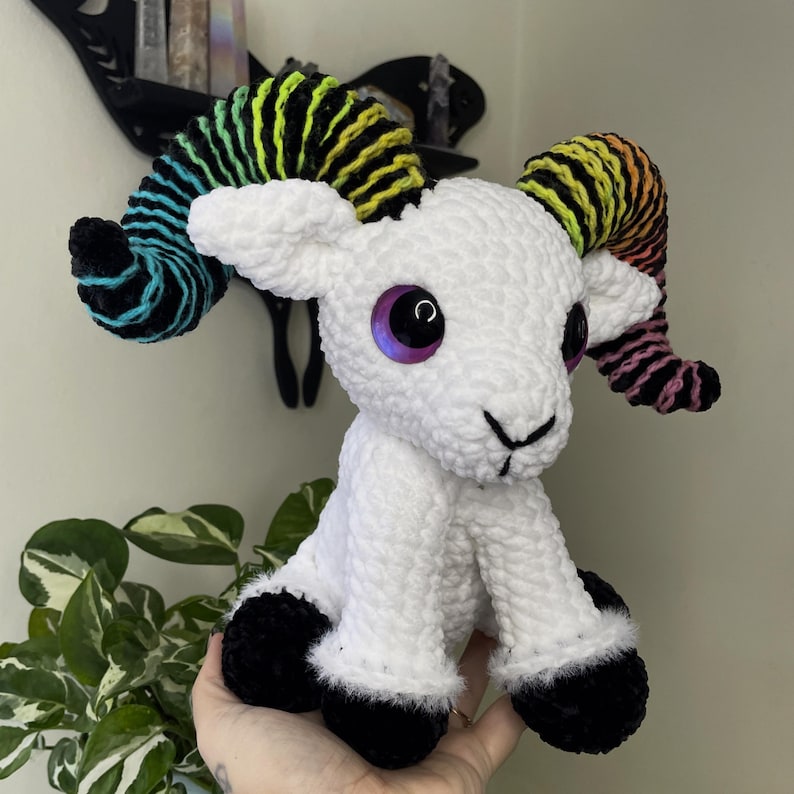 May include: A handmade white crochet ram toy with rainbow horns. The ram has large, purple eyes and black hooves. The plushie is soft and textured, suitable for a nursery or as a unique gift. The horns are striped with various colours.