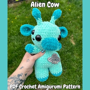 Può includere: Una mucca amigurumi all'uncinetto turchese e verde acqua con grandi occhi neri, un naso blu e una toppa argentata a forma di UFO. Il testo "Alien Cow" e "PDF Crochet Amigurumi Pattern" sono visibili.