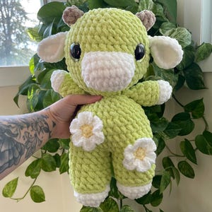 Puede incluir: Una vaca de peluche de crochet verde con detalles blancos y margaritas. La vaca tiene grandes ojos negros y un hocico blanco.