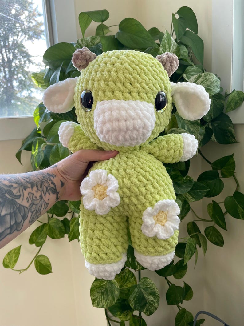 Puede incluir: Una vaca de peluche de crochet verde con detalles blancos y margaritas. La vaca tiene grandes ojos negros y un hocico blanco.