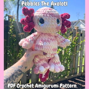 Könnte beinhalten: Ein handgehäkeltes Axolotl-Plüschtier in Rosa und Weiß, mit großen, irisierenden Augen und burgunderfarbenen Kiemen. Der Text "Pebbles The Axolotl" und "PDF Crochet Amigurumi Pattern" sind sichtbar. Das Plüschtier wird vor einem sonnigen Hintergrund im Freien gehalten.