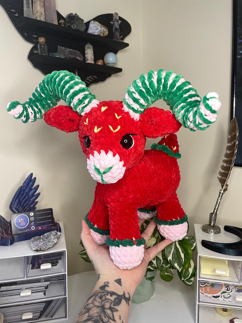 May include: A handmade red plush ram with green and white striped horns. The ram has pink and green accents on its hooves and a white muzzle. The ram is a whimsical, soft sculpture.