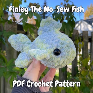 May include: A plush, no-sew fish toy in shades of blue and green, with a black eye. The text "Finley The No-Sew Fish" and "PDF Crochet Pattern" are displayed on the image. The fish has a soft, textured appearance.