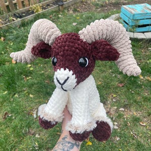 May include: A handmade crochet ram plushie with a burgundy head and hooves, cream-colored body, and tan horns. The ram has black eyes and a white face with a black cross-stitch nose. The plushie is soft and cuddly.