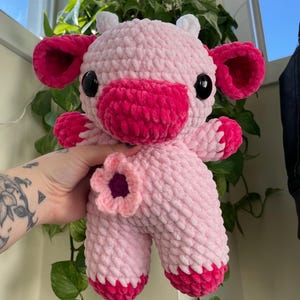 Puede incluir: Una vaca de peluche de crochet rosa y roja con una flor en el costado. La vaca tiene un cuerpo rosa, orejas rojas y una nariz roja.