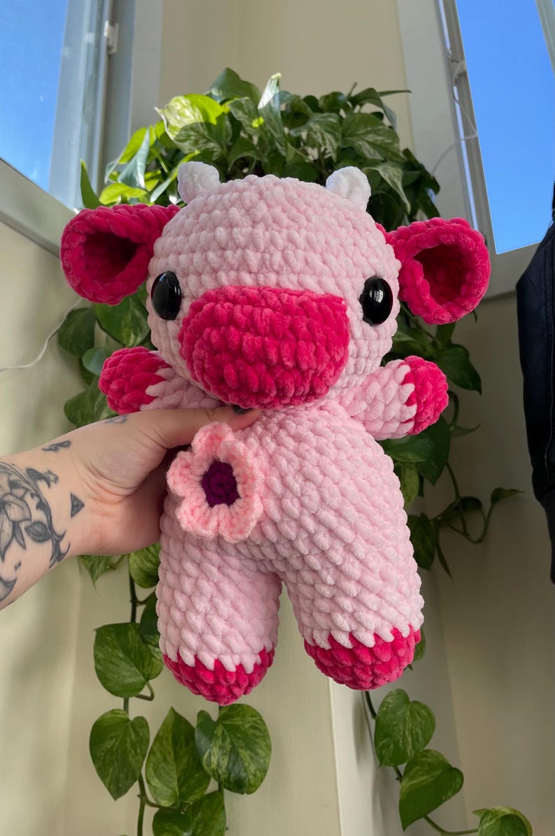 Puede incluir: Una vaca de peluche de crochet rosa y roja con una flor en el costado. La vaca tiene un cuerpo rosa, orejas rojas y una nariz roja.