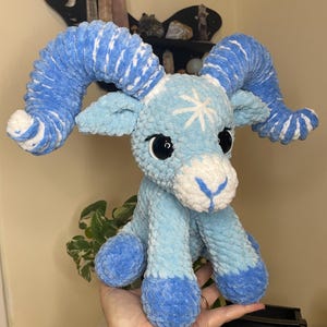 May include: A handmade plush ram in shades of blue and white. The ram has large, curled horns, big black eyes, and a white snowflake design on its forehead. The plushie is made of soft, textured yarn.