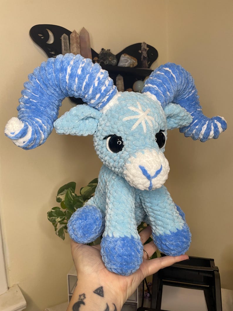 May include: A handmade plush ram in shades of blue and white. The ram has large, curled horns, big black eyes, and a white snowflake design on its forehead. The plushie is made of soft, textured yarn.