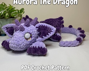 May include: A crocheted purple dragon plush toy named Aurora. The dragon has a long tail and large, sparkly eyes. The toy is lying on a white surface. PDF Crochet Pattern.