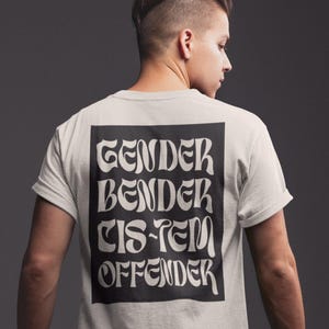 Può includere: T-shirt colorata chiara con le parole "GENDER BENDER CIS-TEM OFFENDER" in nero, con un carattere decorativo, racchiuse in un rettangolo nero. La t-shirt è indossata da una persona, vista da dietro.