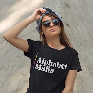 May include: A woman wearing a black t-shirt with the text "Alphabet Mafia" in rainbow colors, black jeans, and sunglasses. She is posing with her hand on her head.