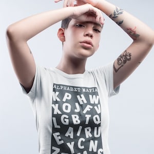 May include: A person wearing a white t-shirt with the text "Alphabet Mafia" and a black graphic of letters. The person is wearing blue jeans and has tattoos on their arms.