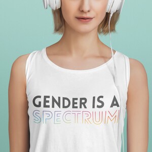 Op de afbeelding: Een persoon draagt een wit tanktop met de tekst "GENDER IS A SPECTRUM" in regenboogkleuren. De persoon draagt een witte koptelefoon en kijkt naar de camera.