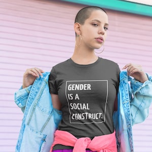 May include: A person wearing a dark gray t-shirt with the text "GENDER IS A SOCIAL CONSTRUCT." printed in white. They are also wearing a light blue denim jacket and a pink and purple skirt.