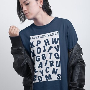 May include: Navy blue t-shirt with the text "ALPHABET MAFIA" in a white rectangle. The shirt features a jumbled arrangement of white letters. The person in the image is wearing a black leather jacket.