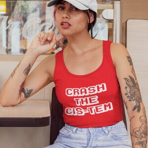May include: A person wearing a red tank top with the text "CRASH THE CIS-TEM" in white pixelated letters. They are wearing a white baseball cap with a rainbow of hearts on the front. The person has tattoos on both arms and is sitting on a wooden chair.