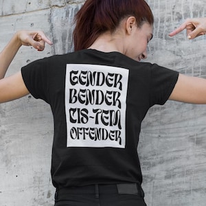 以下が含まれることがあります： 白の様式化されたフォントで「GENDER BENDER CIS-TEM OFFENDER」というテキストが入った黒いTシャツ。シャツは黒いジーンズと組み合わされています。背景は灰色のコンクリートの壁です。