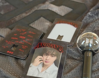 May include: A gray mesh jersey with the number 92 and JIN printed on it, accompanied by two rectangular cards featuring the text ARIRANG, a photo of a person, and a small light stick keychain with a clear dome and a black handle.