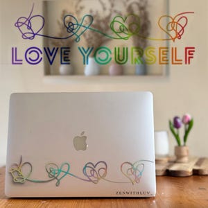 Puede incluir: Un portátil plateado con un diseño de corazón arcoíris y las palabras "LOVE YOURSELF" en colores arcoíris. El diseño se repite en el portátil. El texto "ZENWITHLUV_" está en la parte inferior derecha.