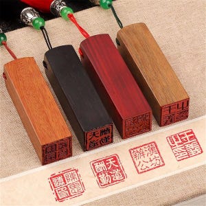 May include: Four rectangular wooden stamps in shades of brown, black, and red. Each stamp has a red ink imprint on the bottom with Chinese characters. The stamps are attached to a red or black cord with a decorative bead.