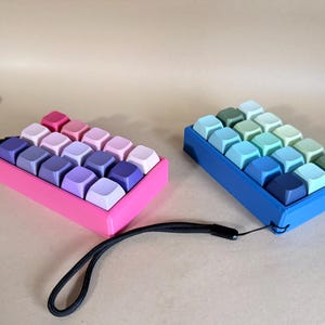 May include: Two miniature keyboard keypads. One is pink with keys in shades of pink and purple. The other is blue with keys in shades of blue and green. Each has a black wrist strap.