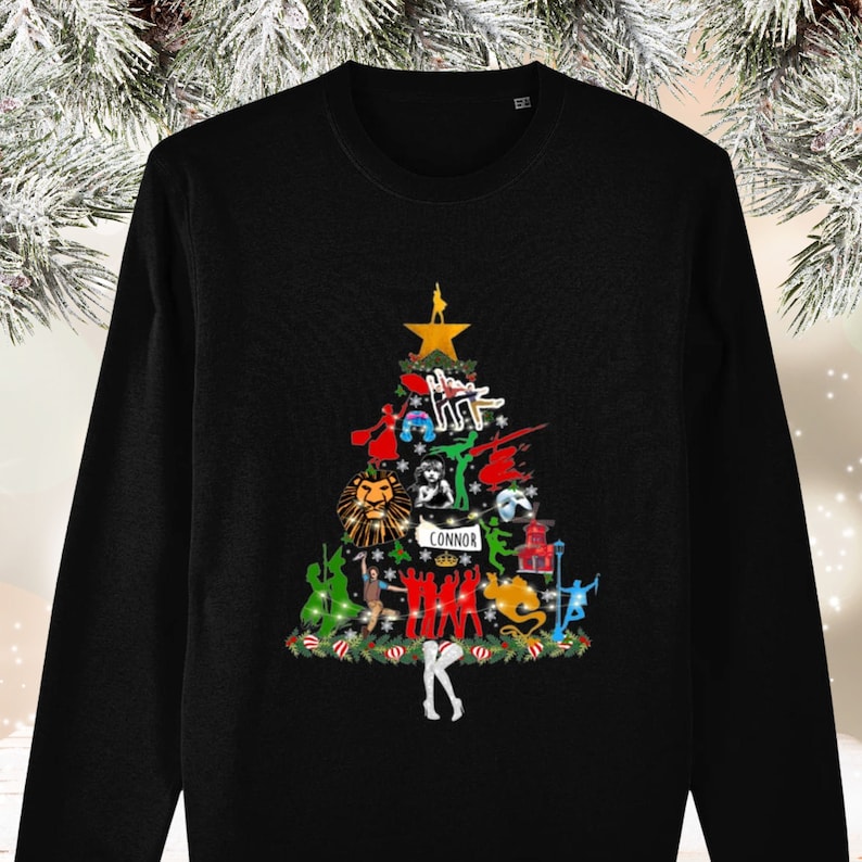 May include: Black long-sleeve shirt with a Christmas tree design featuring characters from popular musicals. The tree is decorated with ornaments and lights. The text "CONNOR" is written on the tree.