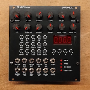 May include: Black electronic music device with knobs, buttons, and ports. The device has the text "BRADSHAW" and "DRUMKID" printed on it. It features a digital display and various input/output jacks. The knobs have red indicators.