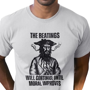 May include: A white t-shirt featuring a black and white illustration of a pirate with the text "THE BEATINGS WILL CONTINUE UNTIL MORAL IMPROVES".