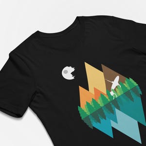 Könnte beinhalten: Ein schwarzes T-Shirt mit einem Grafikdruck einer stilisierten Landschaft mit geometrischen Bergen in Gelb-, Orange- und Brauntönen, einem Wald aus grünen Bäumen, einer blauen Spiegelung und einer weißen Raumstation in der oberen linken Ecke.