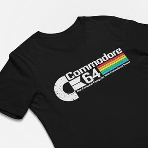 Op de afbeelding: Een zwart t-shirt met korte mouwen voorzien van een verweerd wit Commodore 64-logo met een veelkleurige regenboogstreep en de tekst The Personal Computer With Professional Power in het wit onder het logo.